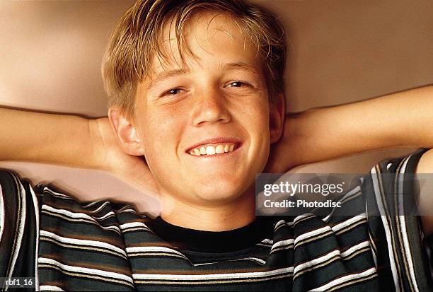 happy older boy with light brown or blonde hair wearing a black and white striped shirt smiles at the camera with his arms raised up and his hands behind his head in a relaxed position. - happy stockfoto's en -beelden