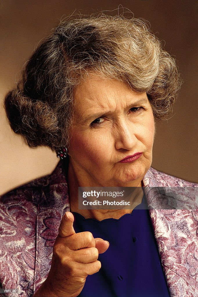 Elderly female woman with shorter graying hair wearing a purple jacket and blue shirt frowns and gives the camera or imaginary person a disapproving look while she shakes her finger and appears to be scolding someone