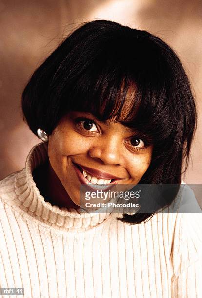 happy young african-american woman female with shorter black hair and beautiful brown eyes wearing a cream turtleneck sweater and earrings smiles and tilts her head - happy stockfoto's en -beelden