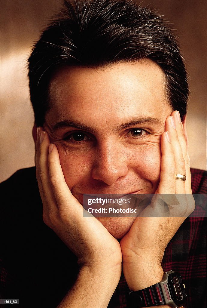 Man male with dark hair wearing a dark shirt rests his chin on his hands and smiles slightly