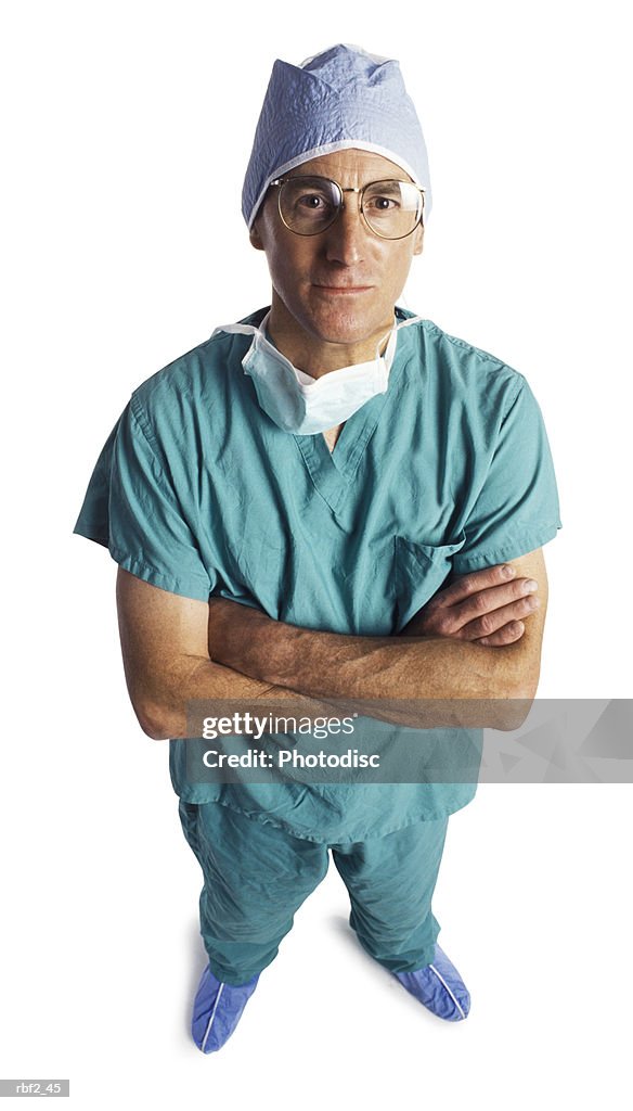 An adult male doctor in scrubs and a surgical mask folds his arms and looks up at the camera