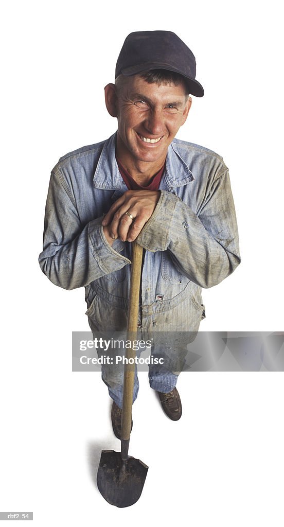 An adult male farmer in work clothes and a shovel smiles up at the camera