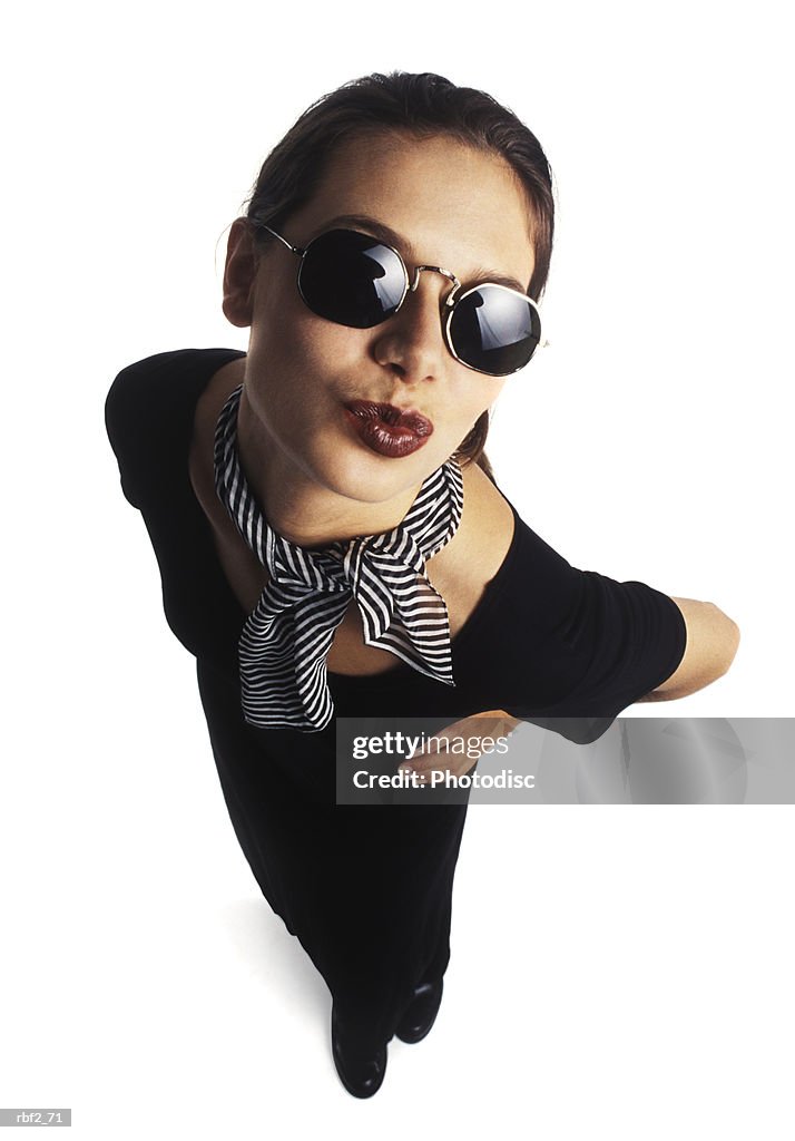 Young adult female model in black dress and sunglasses puckers her lips and kisses up to the camera