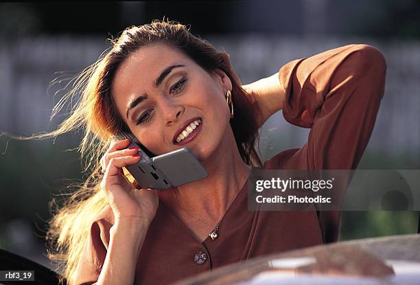 a hispanic woman wearing a brown shirt with a hand behind of head talks on a cellular phone as the sun shines on her hair - hand on head ストックフォトと画像