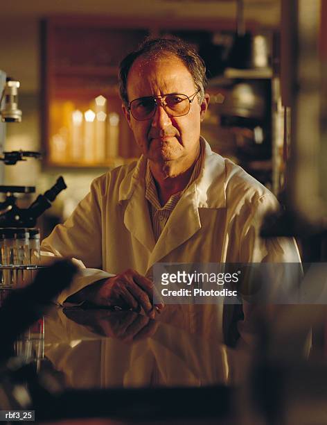 an older scientist wearing a white lab coat sits at a reflective table amongst microscopes - lab coat stock-fotos und bilder