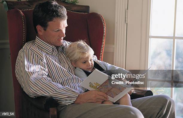 man with tan pants sits in red chair holds his blonde boy in lap reads book in front of glass door - door stockfoto's en -beelden