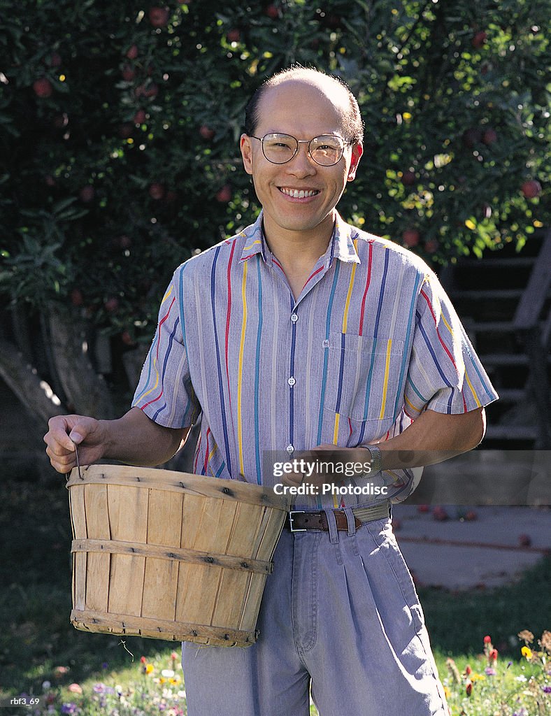 Asian man wears gray pants striped shirt carries a basket smiles standing in a grove of trees