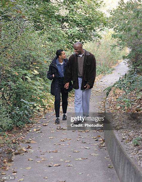 an african-american man wearing denim jeans and a brown coat walks down a green path with an african-american woman wearing black pants and a denim shirt as they hold hands and talk - pants down bildbanksfoton och bilder