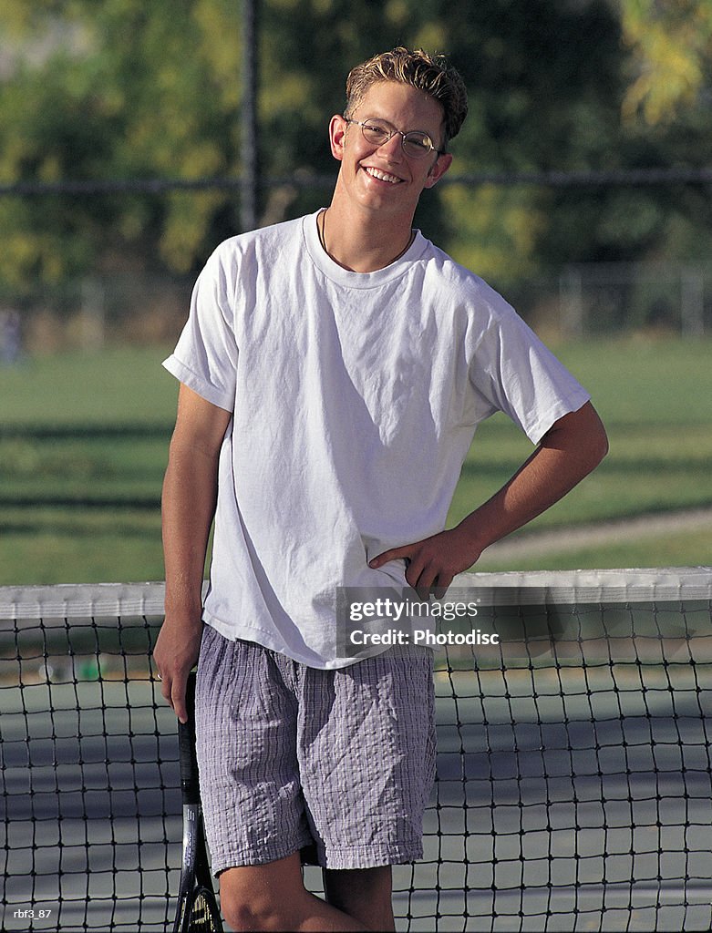 A college-aged man wearing a white shirt and glasses places a hand on his hip as he holds a tennis racket while standing in a tennis court