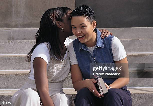 an african-american woman wearing white pants and a white shirt and another african-american woman wearing blue pants and a denim vest whispers in her friend's ear - aother stock pictures, royalty-free photos & images