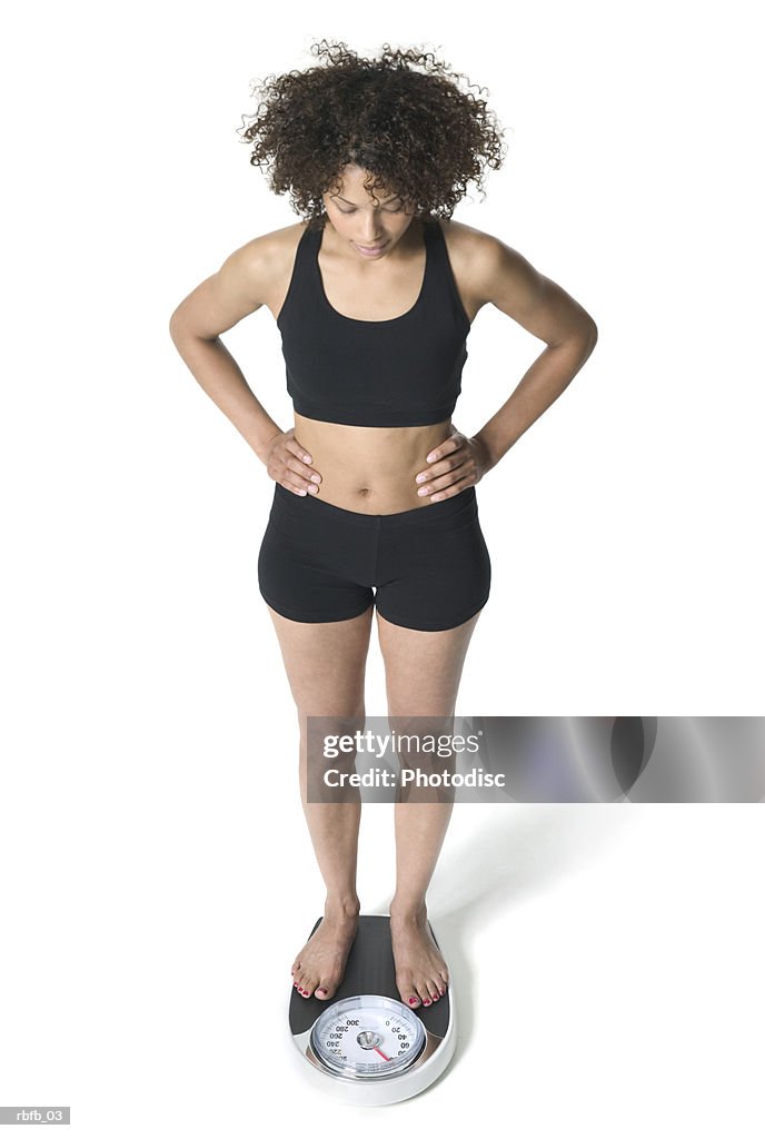 High angle full body of a young adult woman in a black workout outfit as she stands on the scale