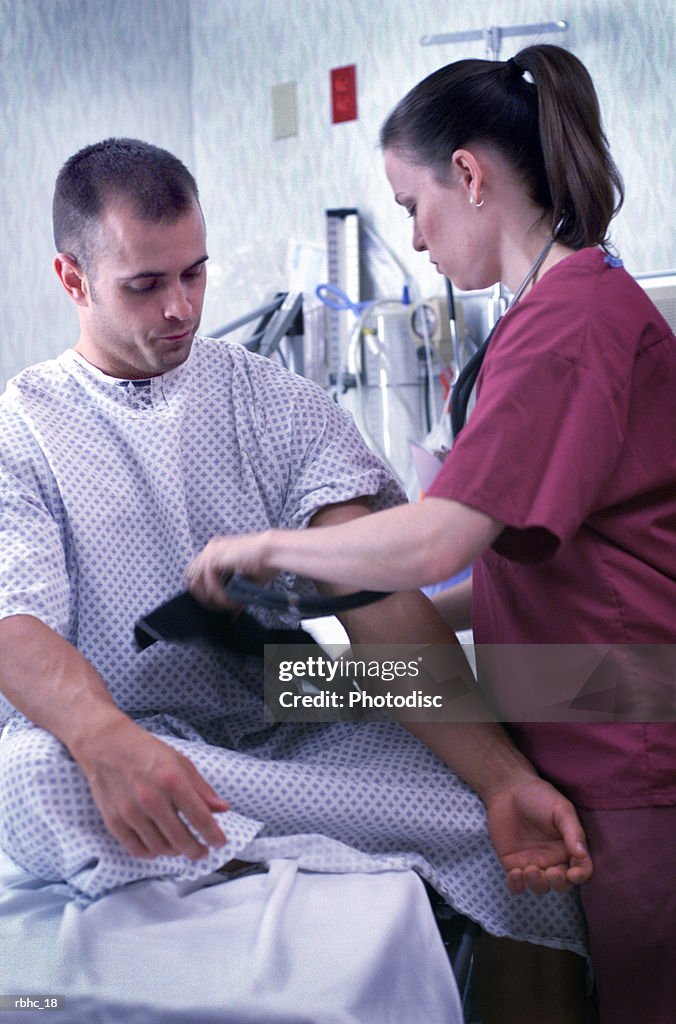 A caucasian nurse in pink scrubs checks the blood pressure of a male caucasian patient in a hopital gown