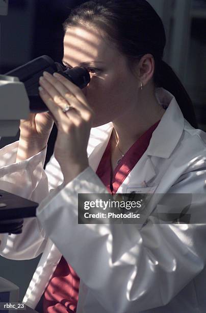 a female caucasian med tech in a white lab coat studies carfully a sample under the microscope - lab coat stock-fotos und bilder