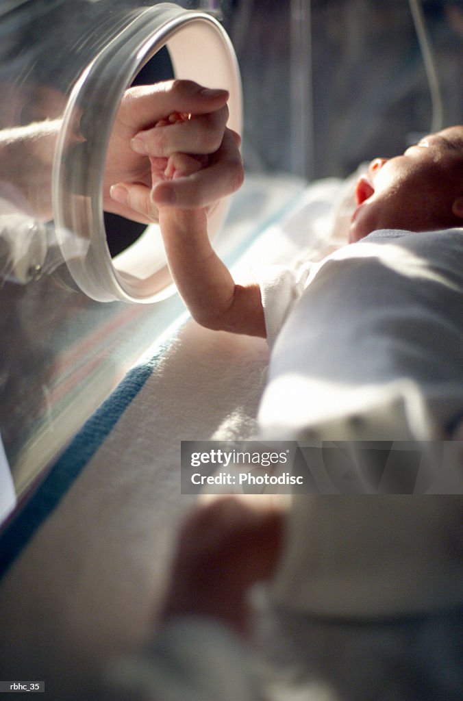 Close up of a caucasain newborn baby laying in an incubator as a nurse takes the baby by the hand