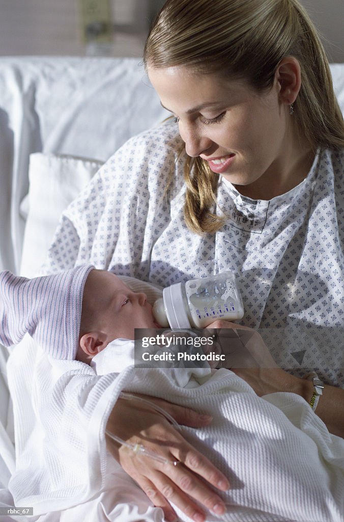 A young caucasian mother sits smiling on a hospital bed as she holds her newborn baby