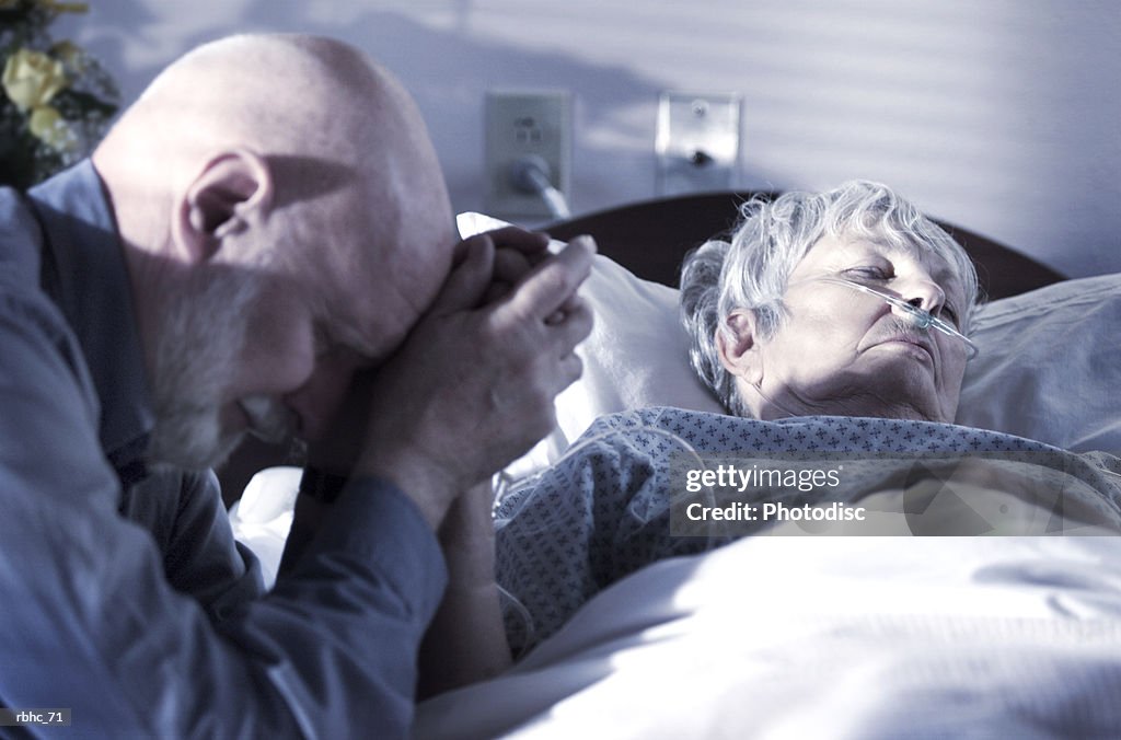 An elderly caucasain gentleman visits and prays over his wife as she tries to recover from an illness