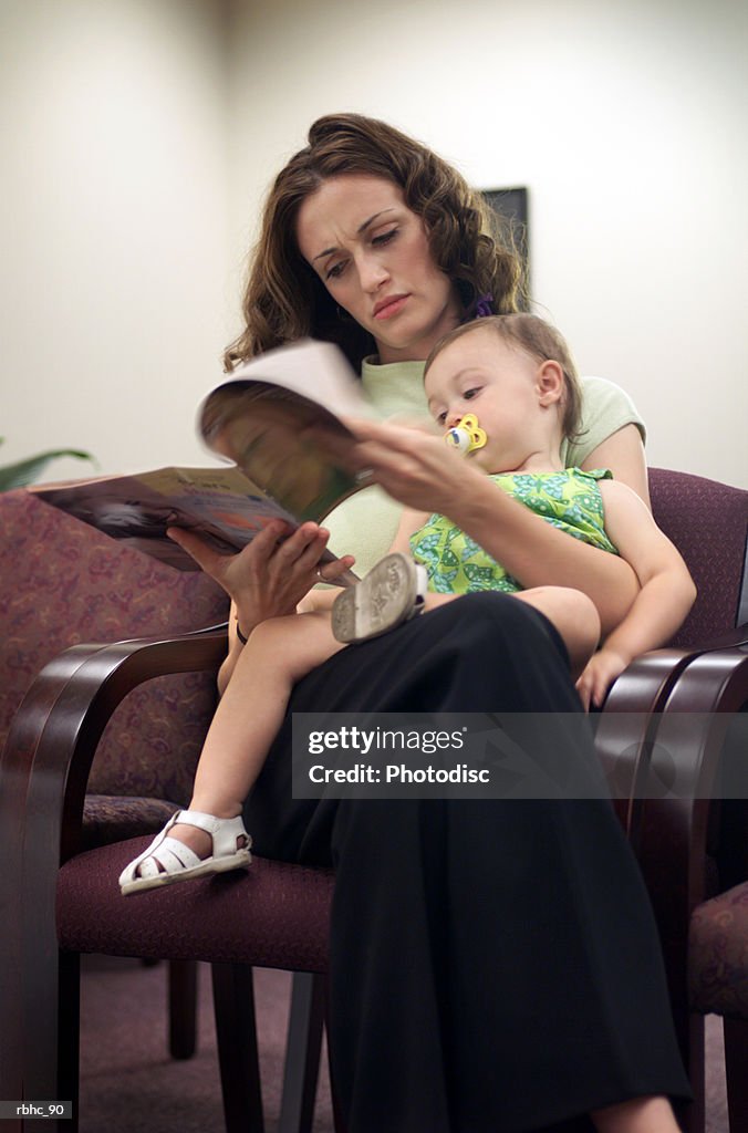 A young caucasian mother sits in a waiting room reading a magazine with her baby on her lap