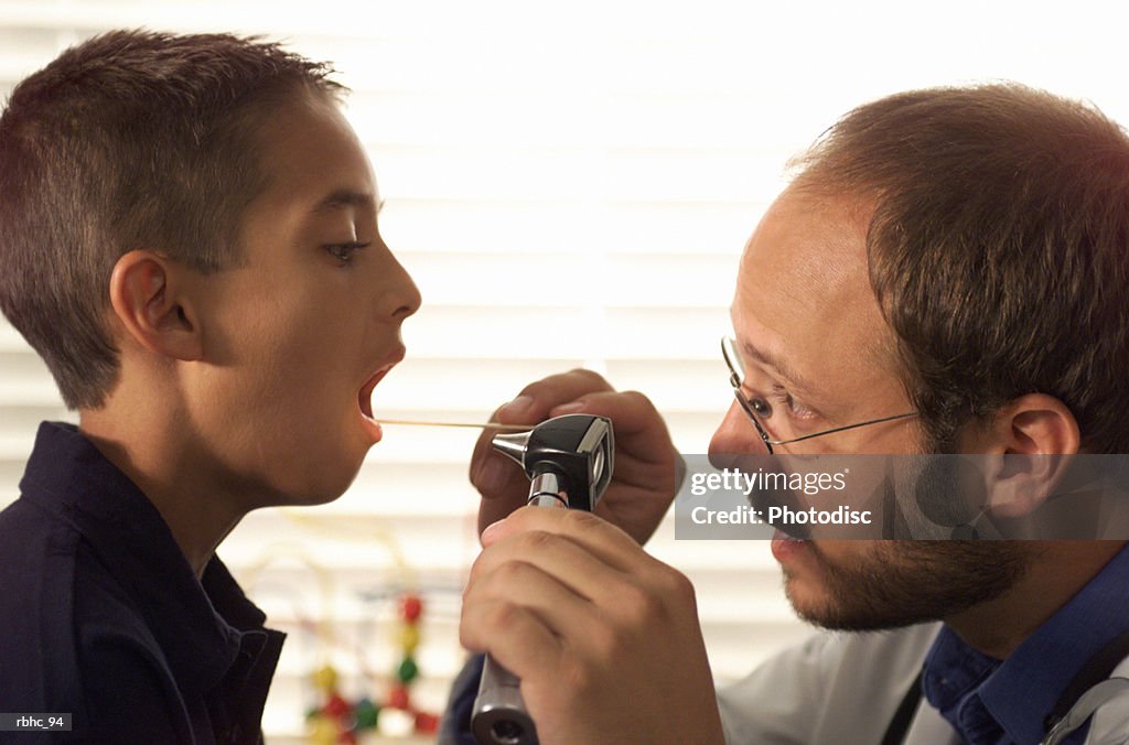 Caucasian male pediatrician with beard examines the throat of a young boy patient during a check up