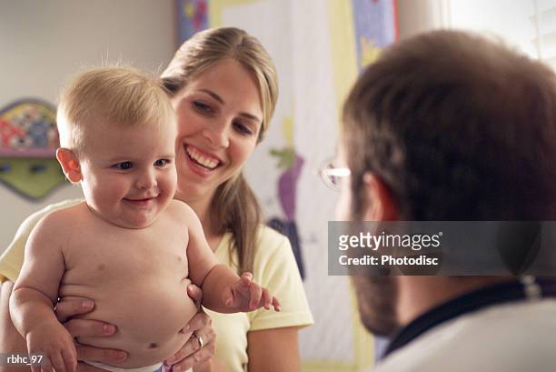 a young caucasian mother sits her baby on her lap as a male pediatrician begins his check up - check up ストックフォトと画像