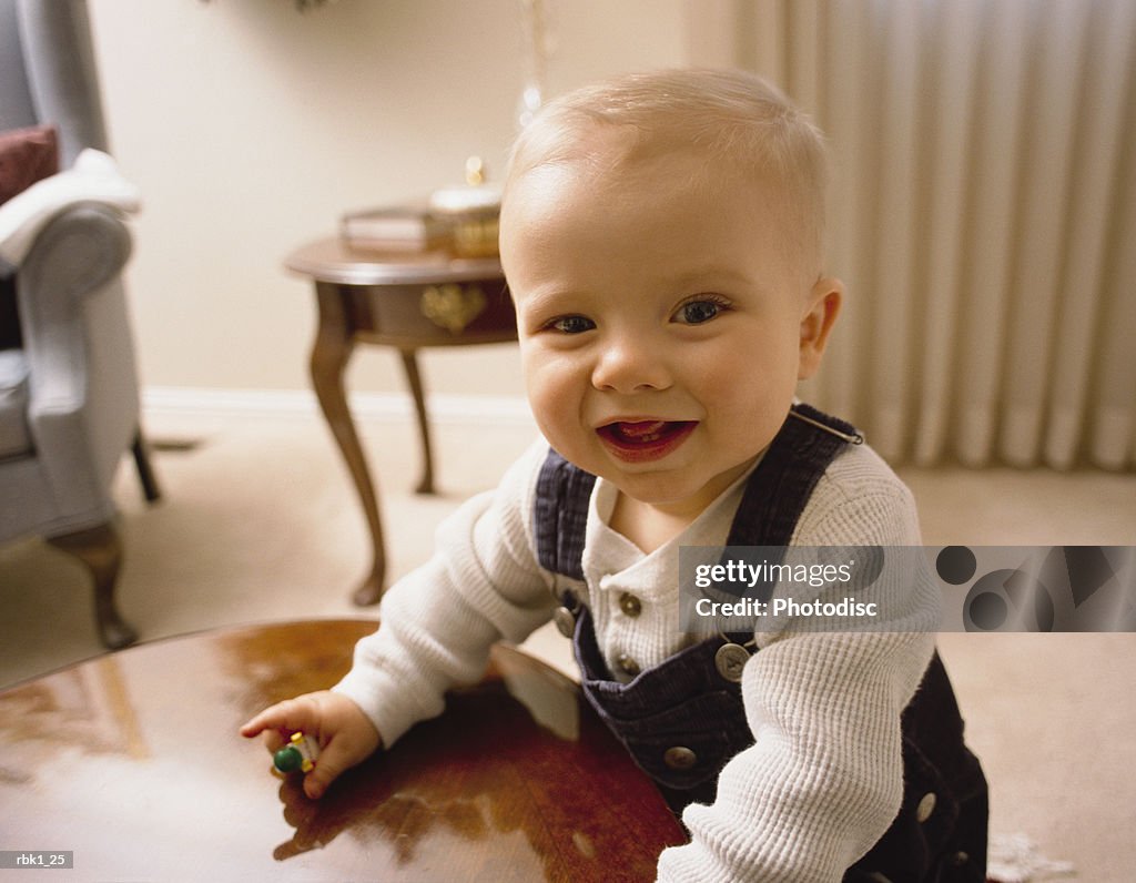 A caucasian blonde male toddler leans against a table and grins