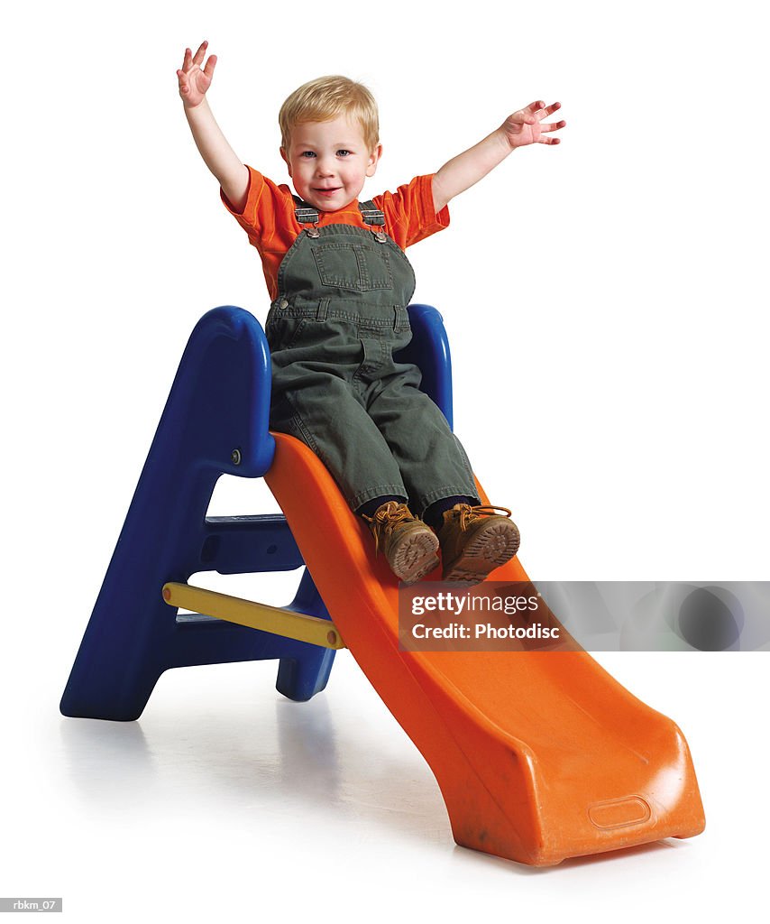 A little caucasian boy throws his arms up as he slides down a toy slide