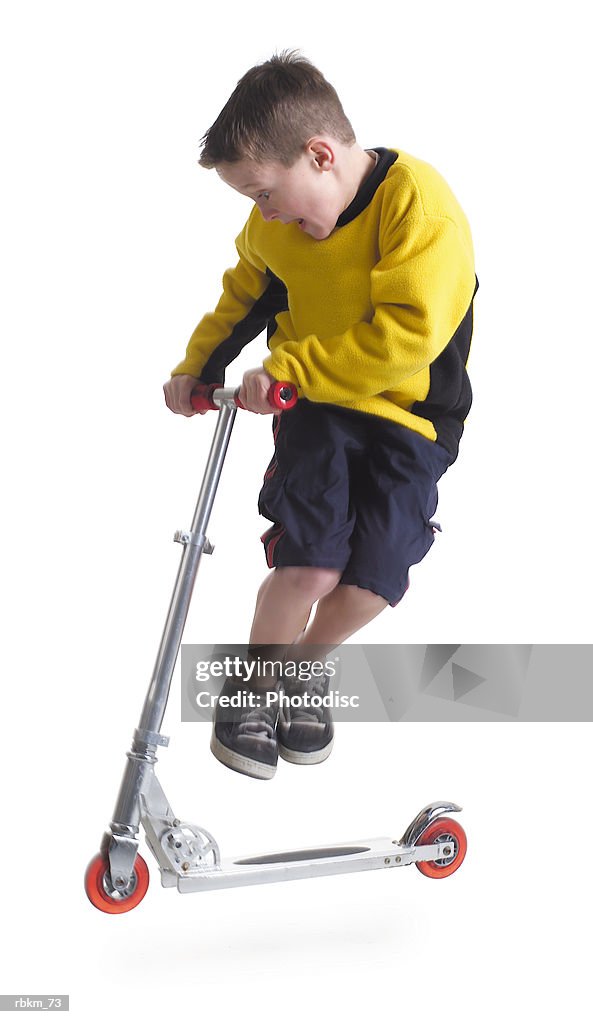 A little caucasian boy in a yellow shirt jumps up while riding his scooter