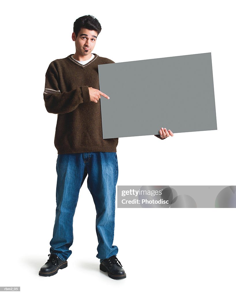 A young ethnic looking man in jeans and a brown sweater points to a large sign by his side