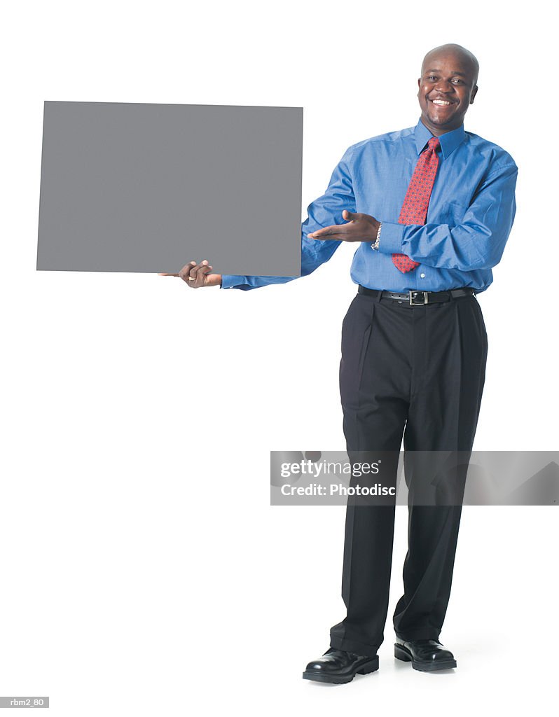 An african american male in black pants and a blue dress shirt holds a blank sign to his side and gestures to it
