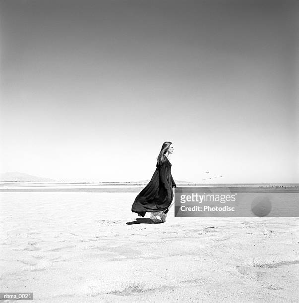 a young white female wearing a long black cloak is walking across the sand at the beach or perhaps the desert but she looks content and peaceful - but stockfoto's en -beelden