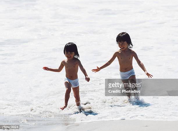 two small asian children in their underwear are running together and playing in the remainder of the ocean waves as they reach the beach - remainder fotografías e imágenes de stock