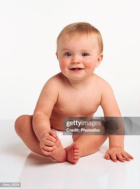 young happy baby with blonde hair blue eyes is sits in diaper smiles to camera grasping her toes - baby blue eyes stock pictures, royalty-free photos & images