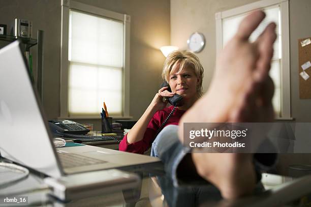 a blonde woman leans back in her chair while on the phone and puts her bare feet on the desk - legs crossed at ankle stock pictures, royalty-free photos & images
