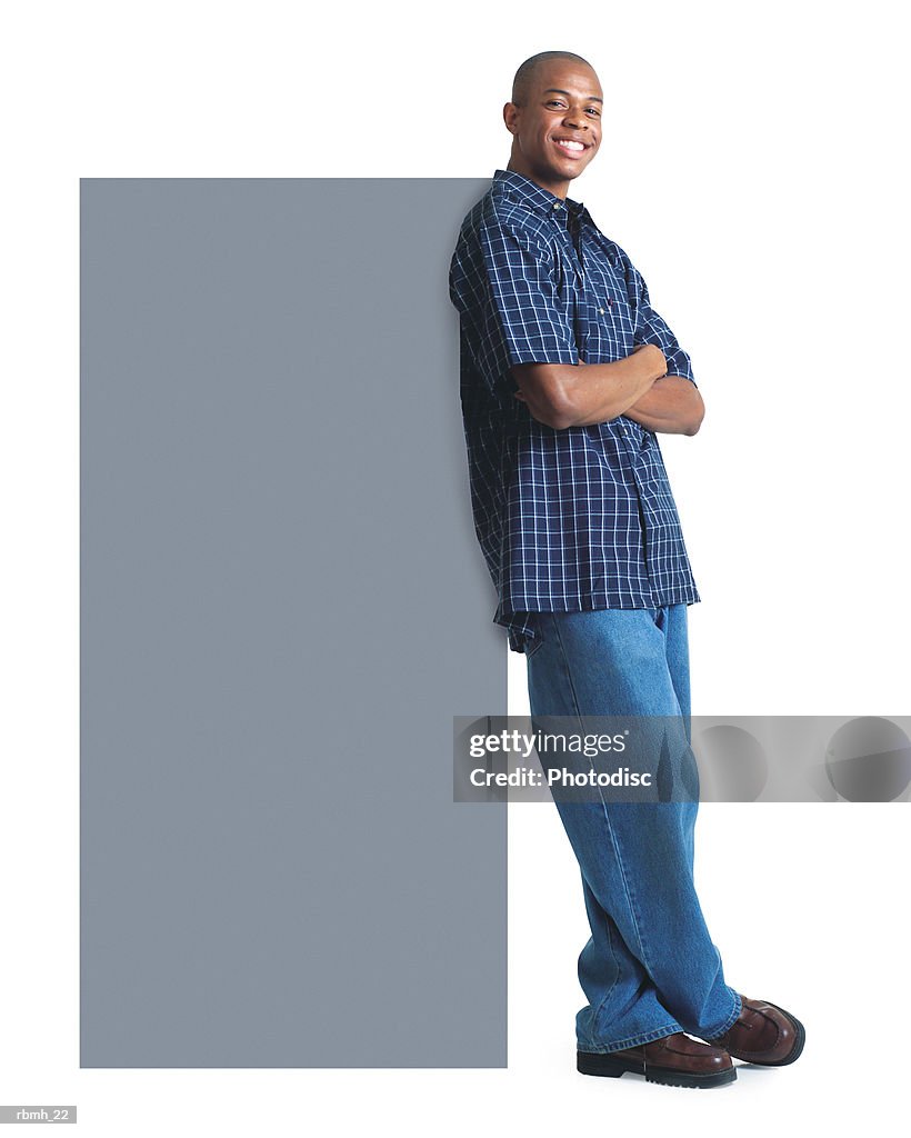 A young african american man in jeans and a plaid shirt smiles as he leans against a large blank sign