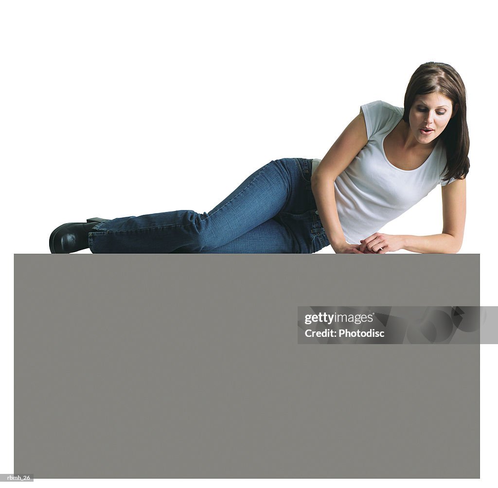 A young attractive caucasian woman in jeans and a white t-shirt lays on a large blank sign and looks down at it in amusement as she props herself up on her elbow