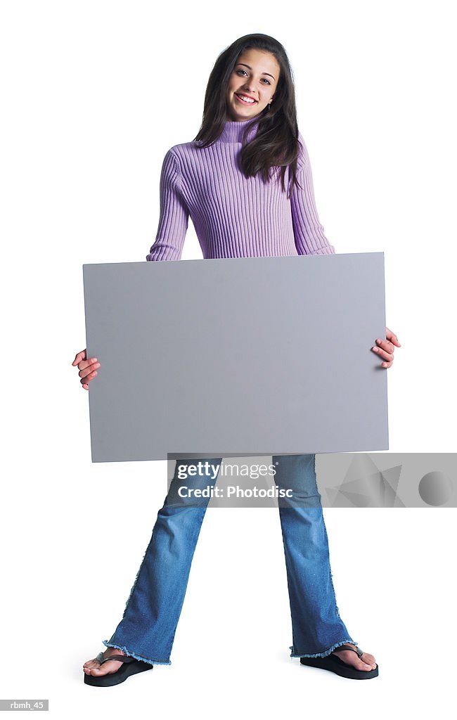 A cute ethnic female wearing jeans and a purple shirt stands with her feet shoulder width apart and holds a blank sign in front of her and smiles