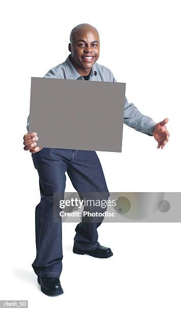 a young bald african american man wearing dark slacks and a grey shirt steps forward with one hand holding a blank sign and one hand out to the side while smiling - bald stock-fotos und bilder