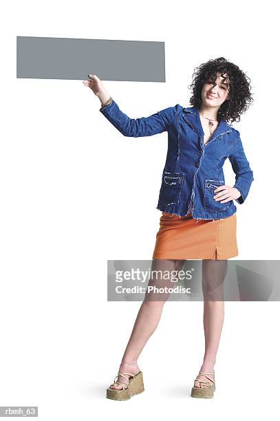 a caucasian girl with brown curly hair wearing a denim shirt and an orang skirt stands with a hand on one of her hips and her other hand holding a blank sign up to the side of her head - hand on head ストックフォトと画像