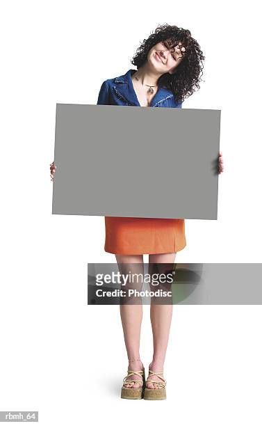 a caucasian girl with brown curly hair wearing a denim shirt and an orang skirt holds a blank sign in front of her with both hands and her head tilted to one side as she smiles - curly stock pictures, royalty-free photos & images
