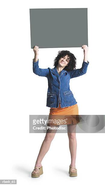a caucasian girl with brown curly hair wearing a denim shirt and an orang skirt holds a blank sign above her head with both hands as she tilts her head and smiles - curly stock pictures, royalty-free photos & images