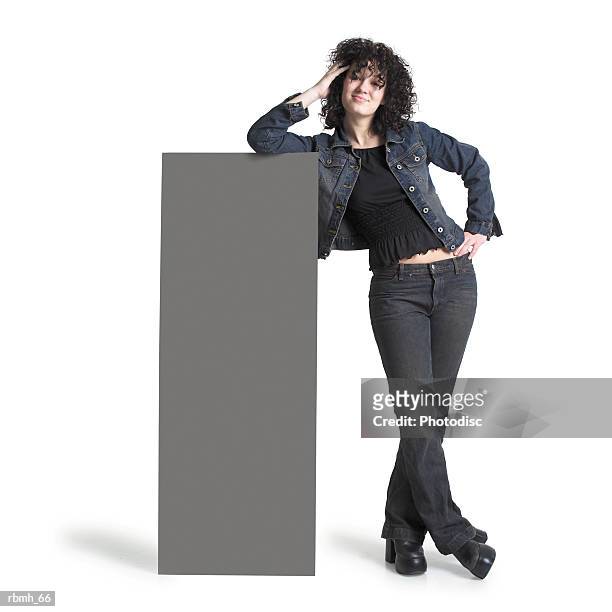 a caucasian girl with brown curly hair wearing jeans a black shirt and a jean jacket stands next to a large blank sign and leans on it with one arm and she has her other arm resting on her hip while she smiles at the camera - next imagens e fotografias de stock