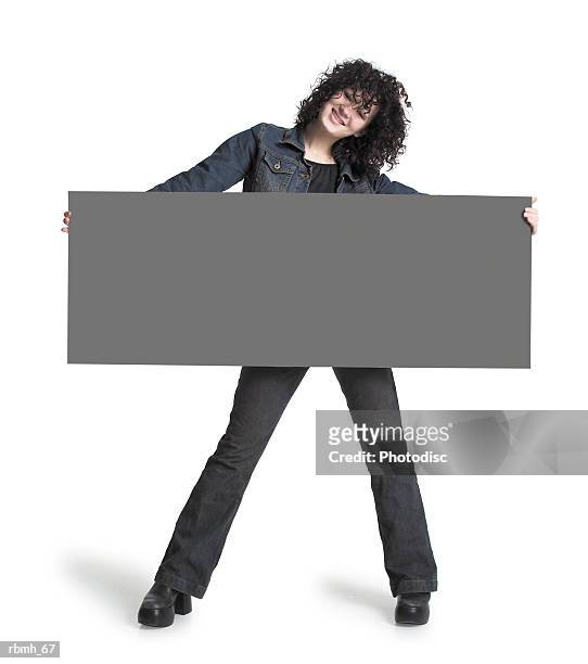 a caucasian girl with brown curly hair wearing jeans a black shirt and a jean jacket holds a large blank sign out in front of her body as she tosses her head to one side and smiles - curly stock pictures, royalty-free photos & images