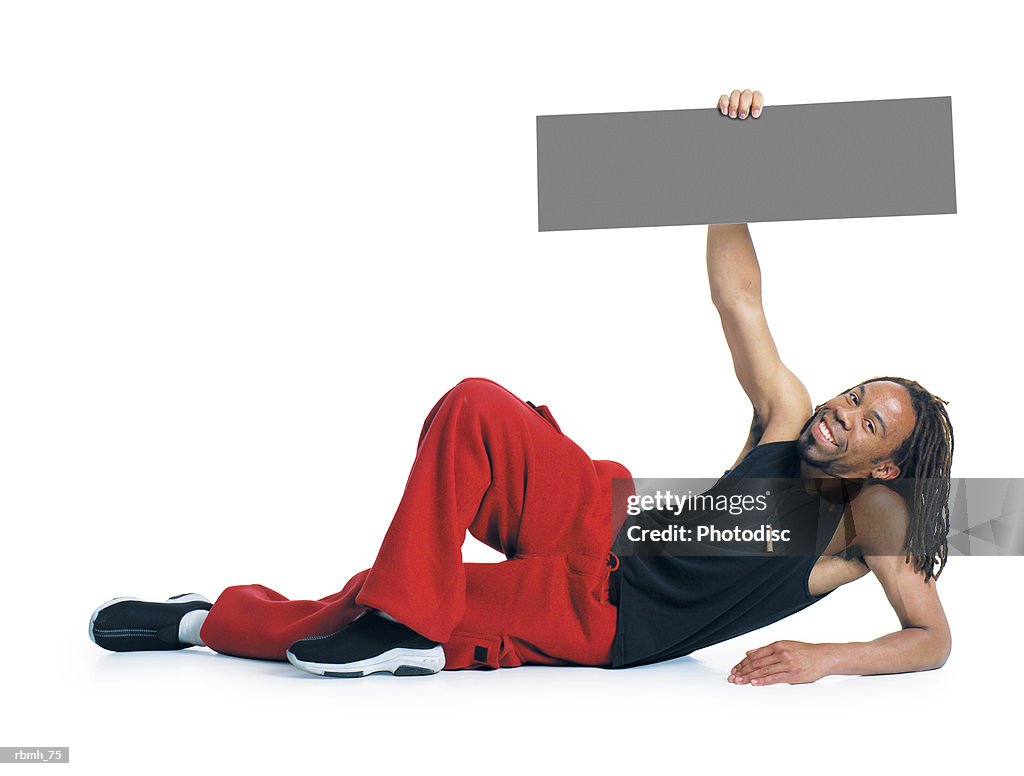 An african american man with dreadlocks wearing red pants and a black tank top lays on the ground propped up on one elbow while his other arm holds a blank sign above him and he smiles