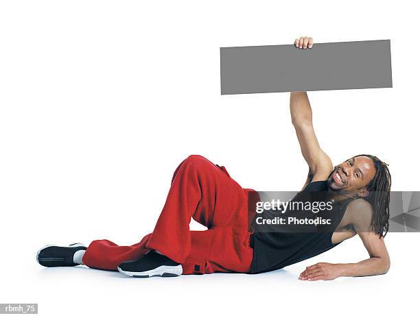 an african american man with dreadlocks wearing red pants and a black tank top lays on the ground propped up on one elbow while his other arm holds a blank sign above him and he smiles - propped stock pictures, royalty-free photos & images