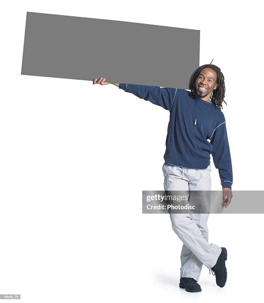 An african american man with dreadlocks wearing kakhi pants and a blue shirt holds a blank sign with one hand and rests it on his shoulder as he stands with his legs crossed