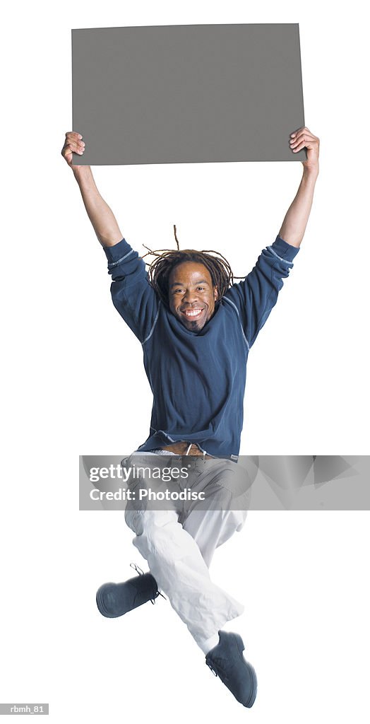An african american man with dreadlocks wearing kakhi pants and a blue shirt jumps in the air with is legs crossing as he holds a blank sign above his head with two hands