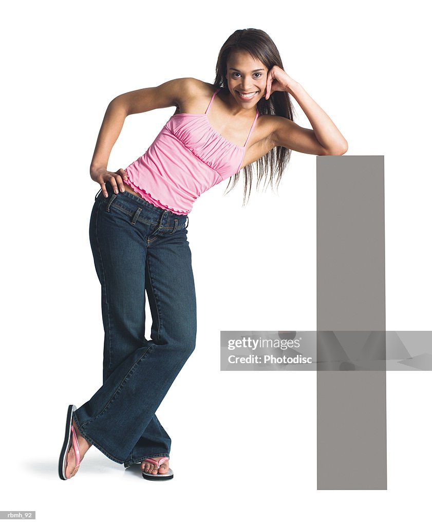 A young attractive african american girl in jeans and a  pink tank top stands with her legs crossed and leans on a blank sign propping her head up on her elbow