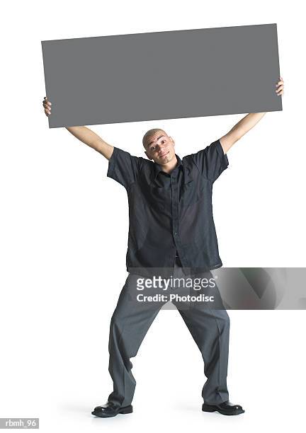 a young hispanic male wearing grey slacks and a black shirt stands with his feet shoulder width apart and his knees bent and head cocked while he spreads his arms overhead to hold a blank sign - width stock pictures, royalty-free photos & images