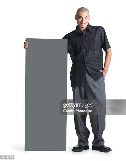 a young hispanic male wearing grey slacks and a black shirt stands next to a blank sign and wraps his arm around the top as he smiles - next stockfoto's en -beelden