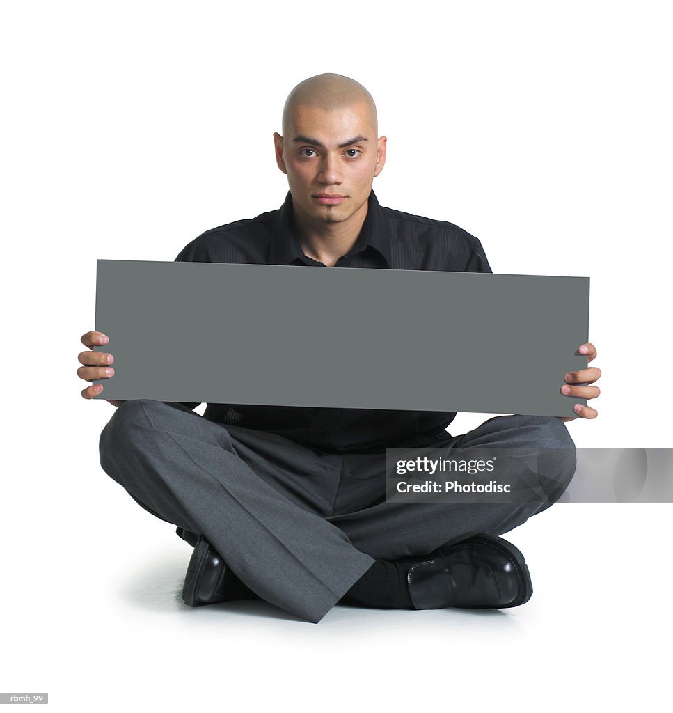 A young hispanic male in grey slacks and a black shirt sits cross-legged on the floor as he holds a blank sign on his lap with both hands and looks straight ahead at the camera