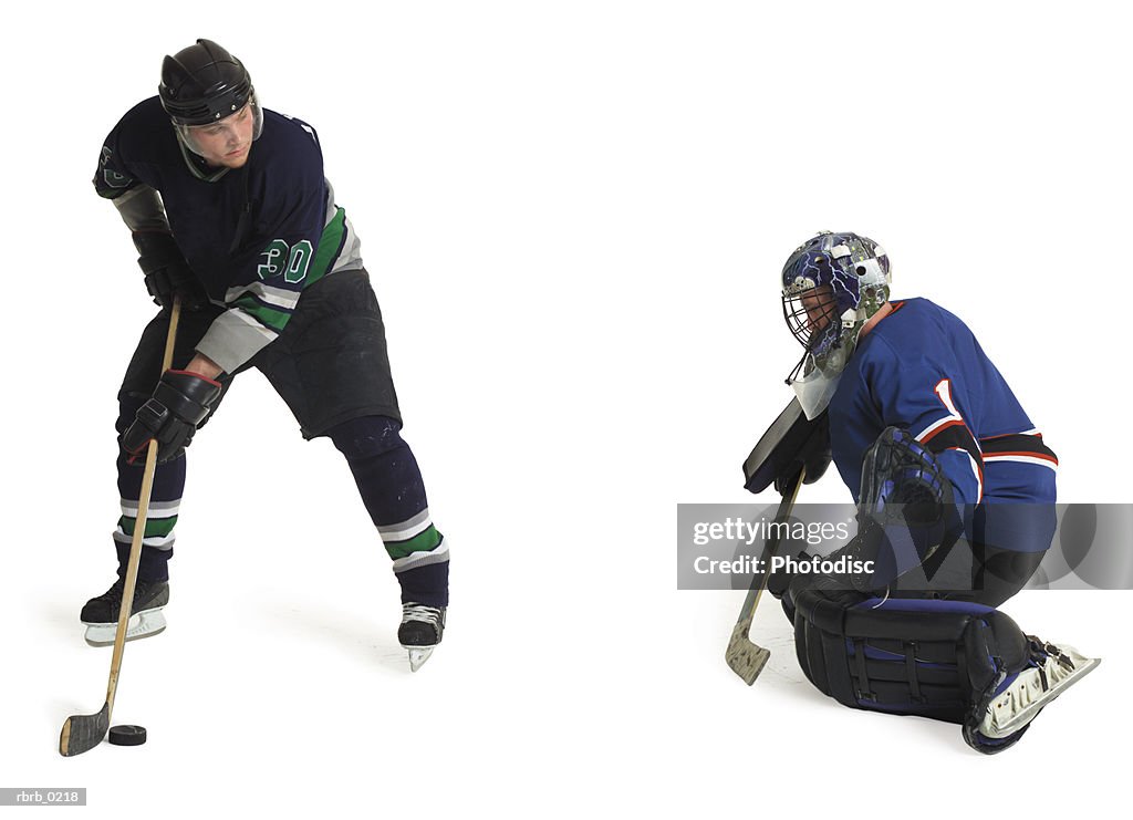 Two adult caucasian male hockey players from opposing teams play as one prepares to shoot and goalie crouches and blocks